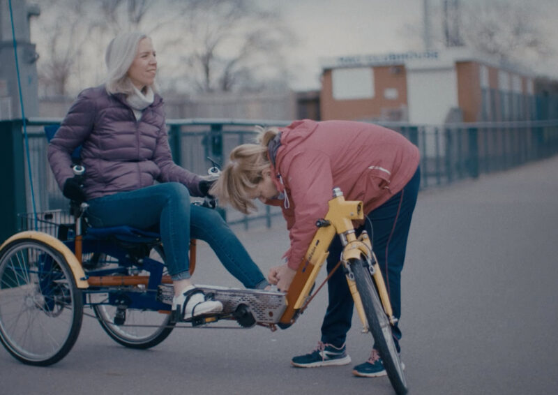 Lady sitting on a special bicycle with another lady buckling her foot - Mindset Science bicycle